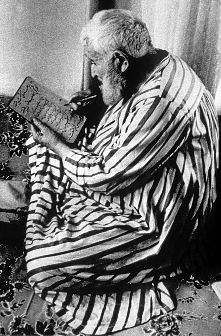 Necmeddin Okyay’ı geleneğe bağlı oturuşla dizi üstünde yazarken gösteren bir fotoğraf (M. Uğur Derman fotoğraf arşivi)